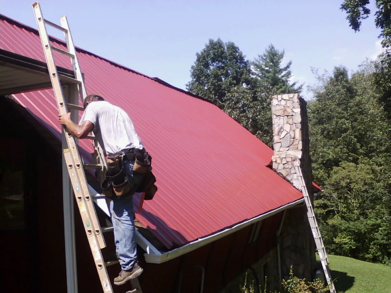 Roofer on a ladder beside a red metal roof and stone chimney — Soffit & Fascia Repair by Roofing Brothers in Rapidan